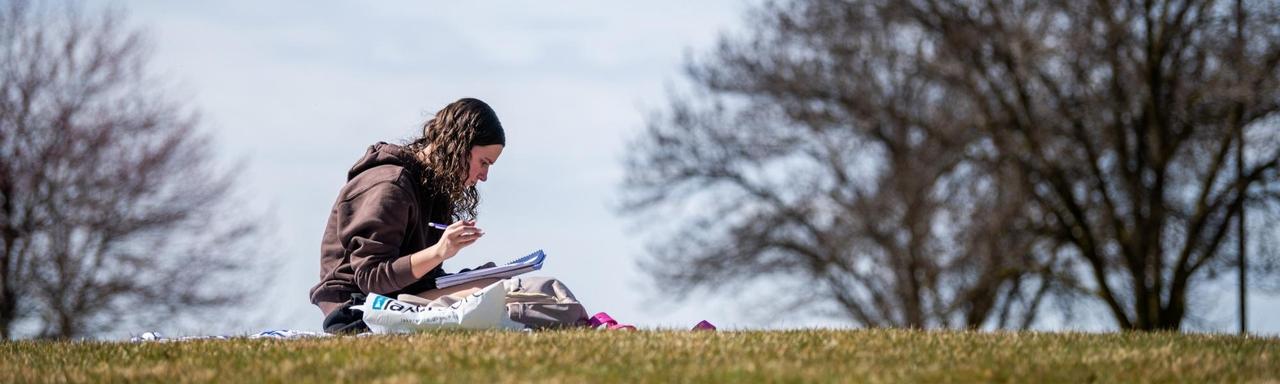 Students studies on a grassy hill with trees in the background